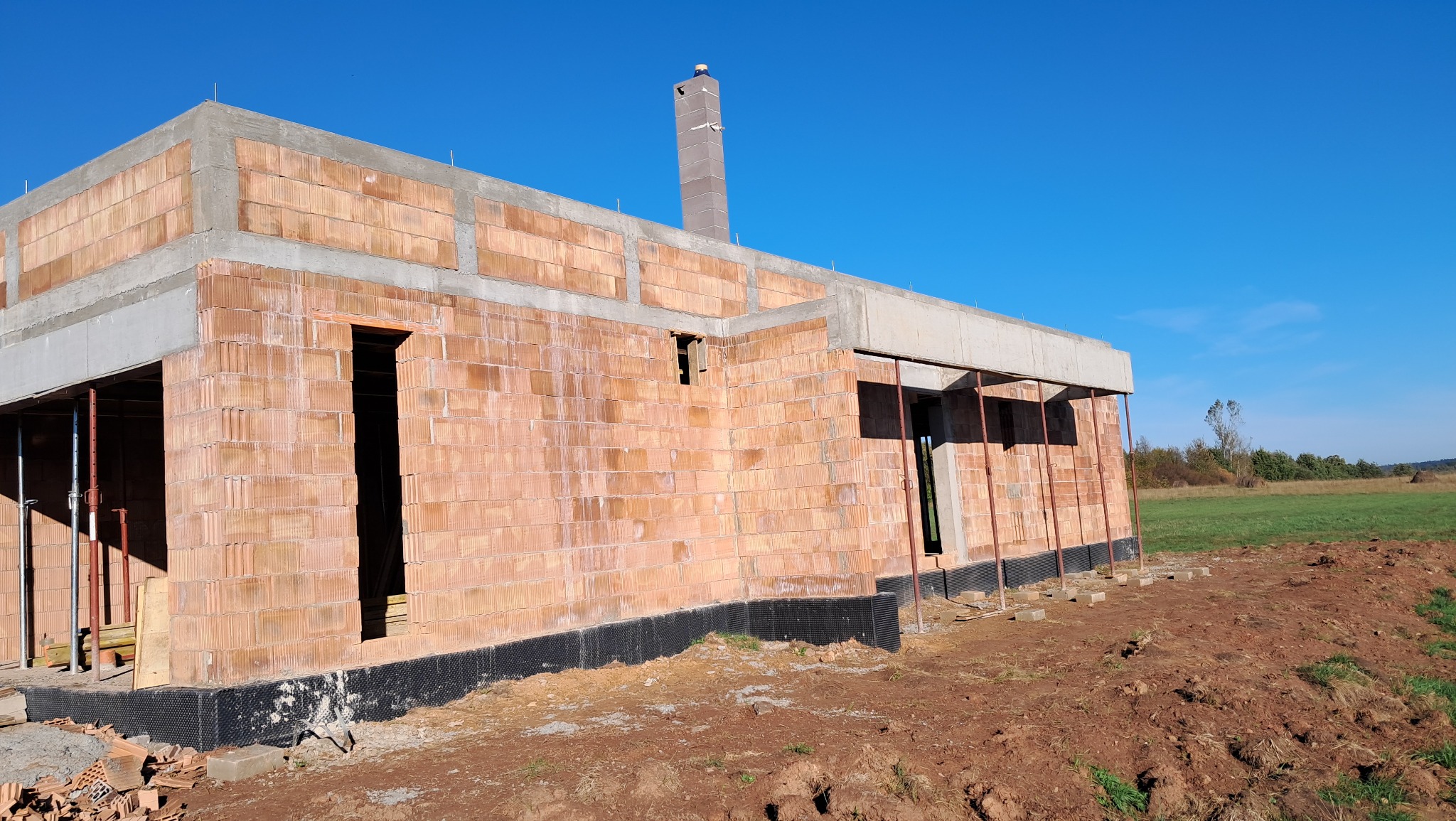 Surowy, niewykończony budynek z cegły, z widoczną izolacją fundamentów i kominem, na tle błękitnego nieba i zielonego krajobrazu.