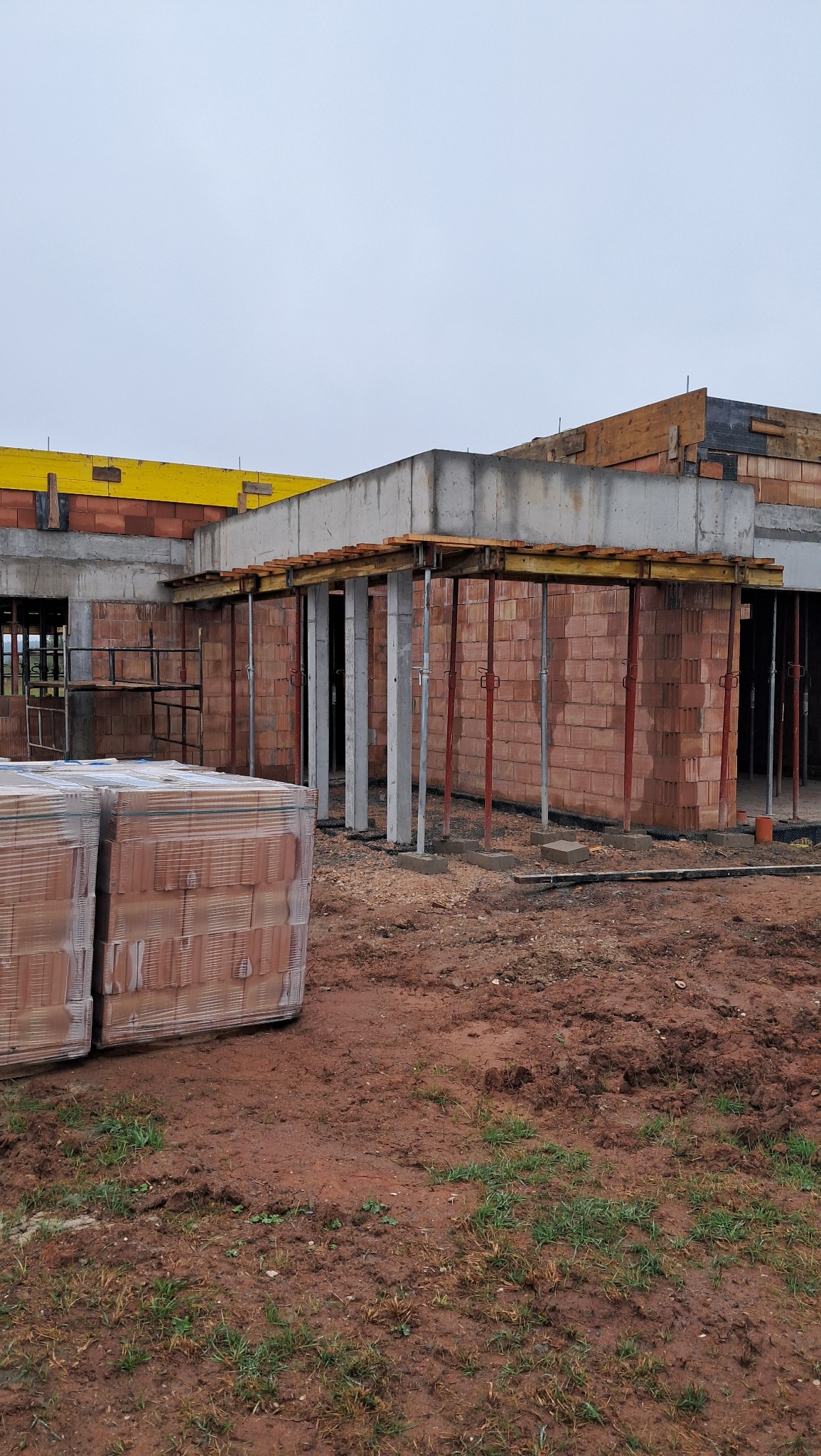 Budowa domu murowanego w trakcie realizacji: widoczne ściany z czerwonej cegły, podpory stropu, rusztowania i palety cegieł na błotnistym terenie.