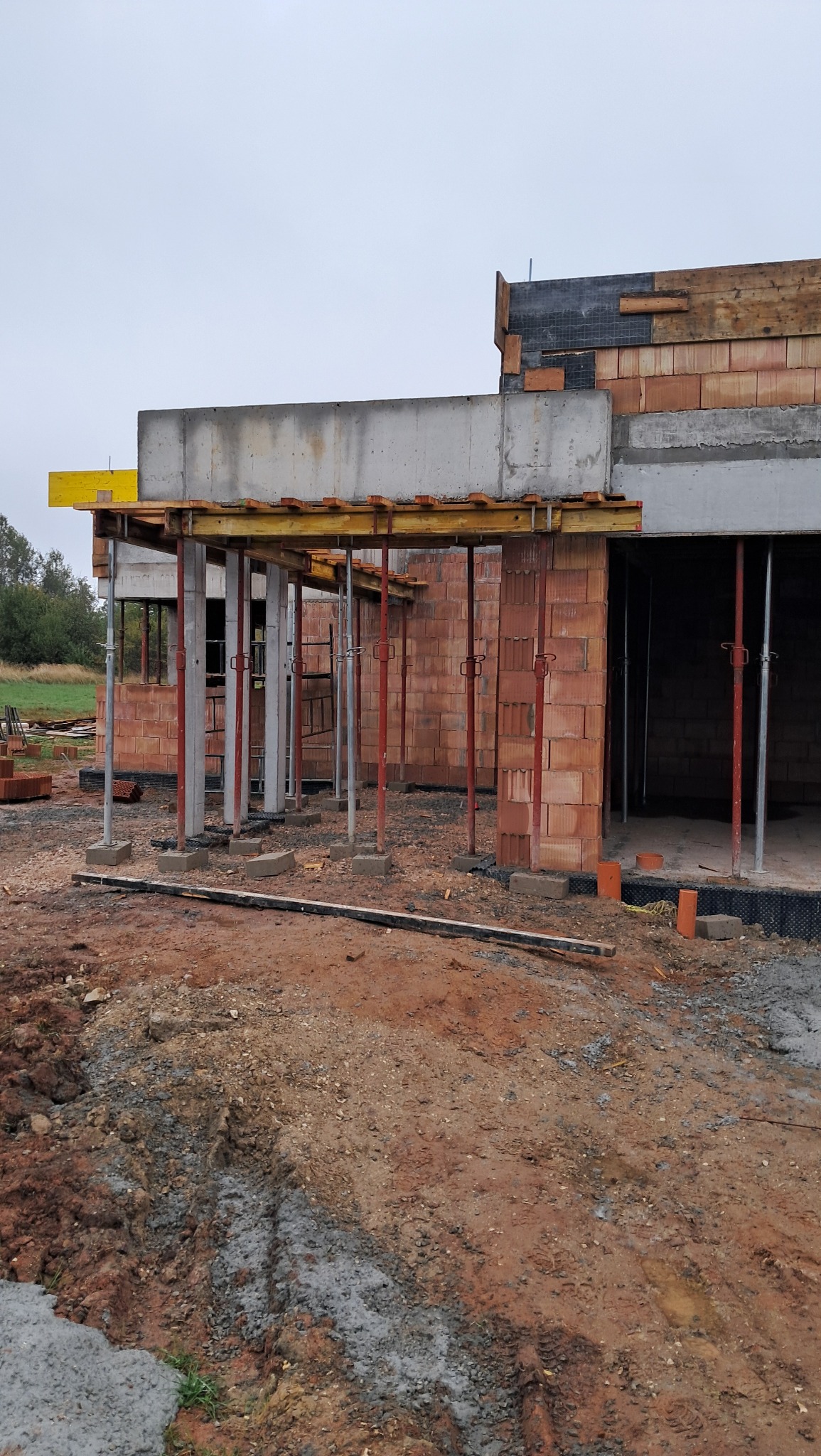 Wznoszenie ścian z czerwonej cegły na budowie domu, widoczne podpory i szalunki stropu, ziemiste podłoże.