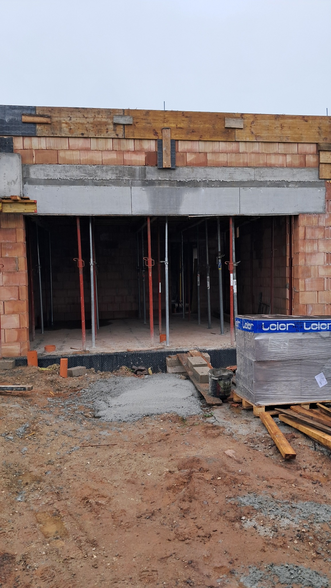 Budowa domu z czerwonej cegły w Bliżynie, widoczne podpory stalowe, paleta z materiałami budowlanymi oraz drewniane elementy szalunku.