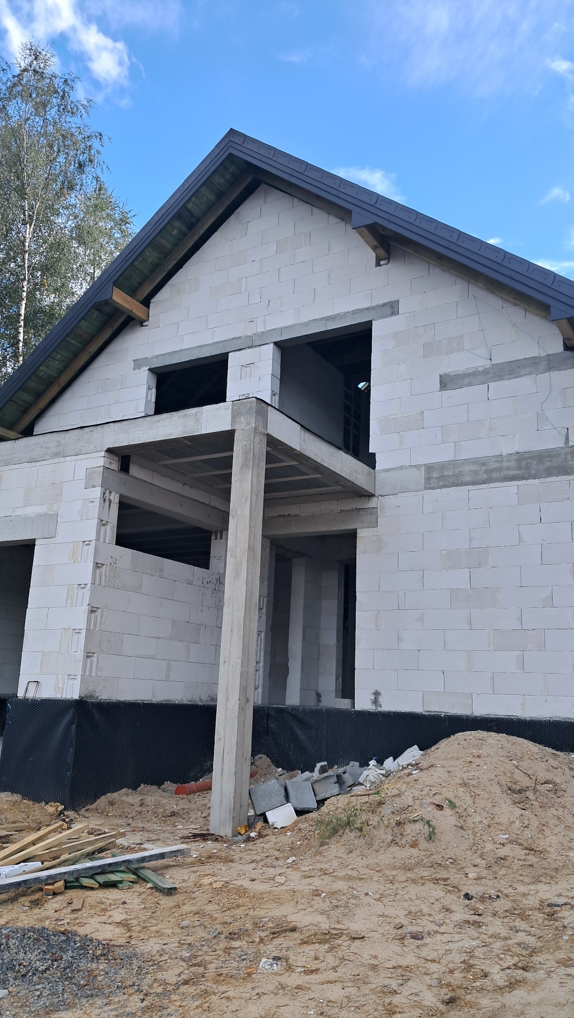 Surowy stan domu w trakcie budowy, ściany z białych bloczków, widoczne otwory okienne i drzwiowe, betonowe filary podtrzymujące strop, czarna izolacja fundamentów, ziemia i gruz przed budynkiem...