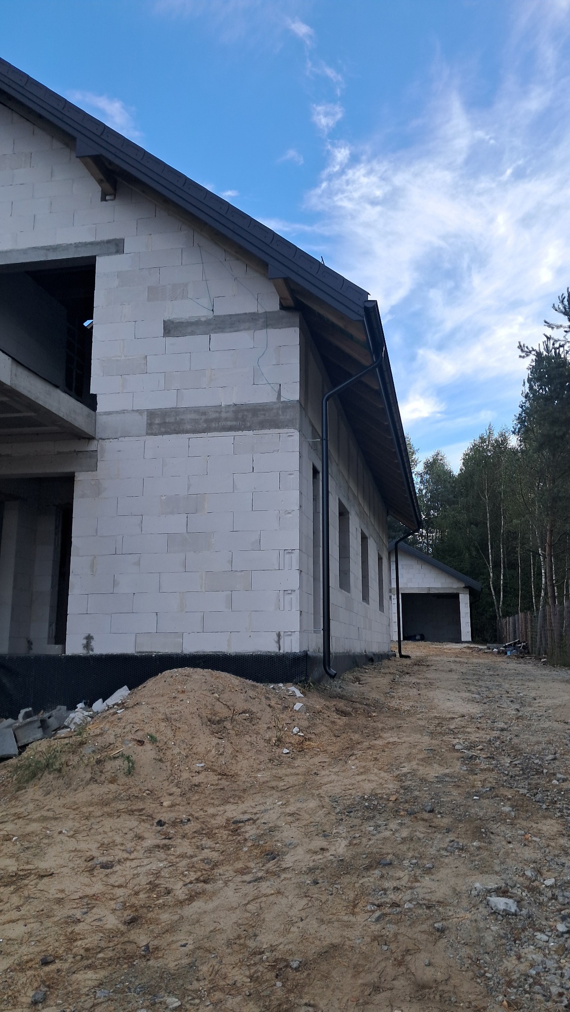 Surowy stan domu z białych bloczków, widoczne rynny, fundament zabezpieczony czarną folią, na pierwszym planie hałda piasku, w tle budynek gospodarczy i drzewa.