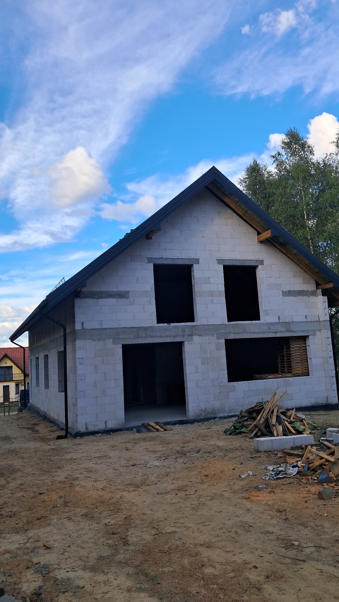 Surowy stan domu z białego bloczka, widoczne otwory okienne i drzwiowe, czarne orynnowanie, niezagospodarowany teren budowy, częściowo widoczne palety wewnątrz budynku.