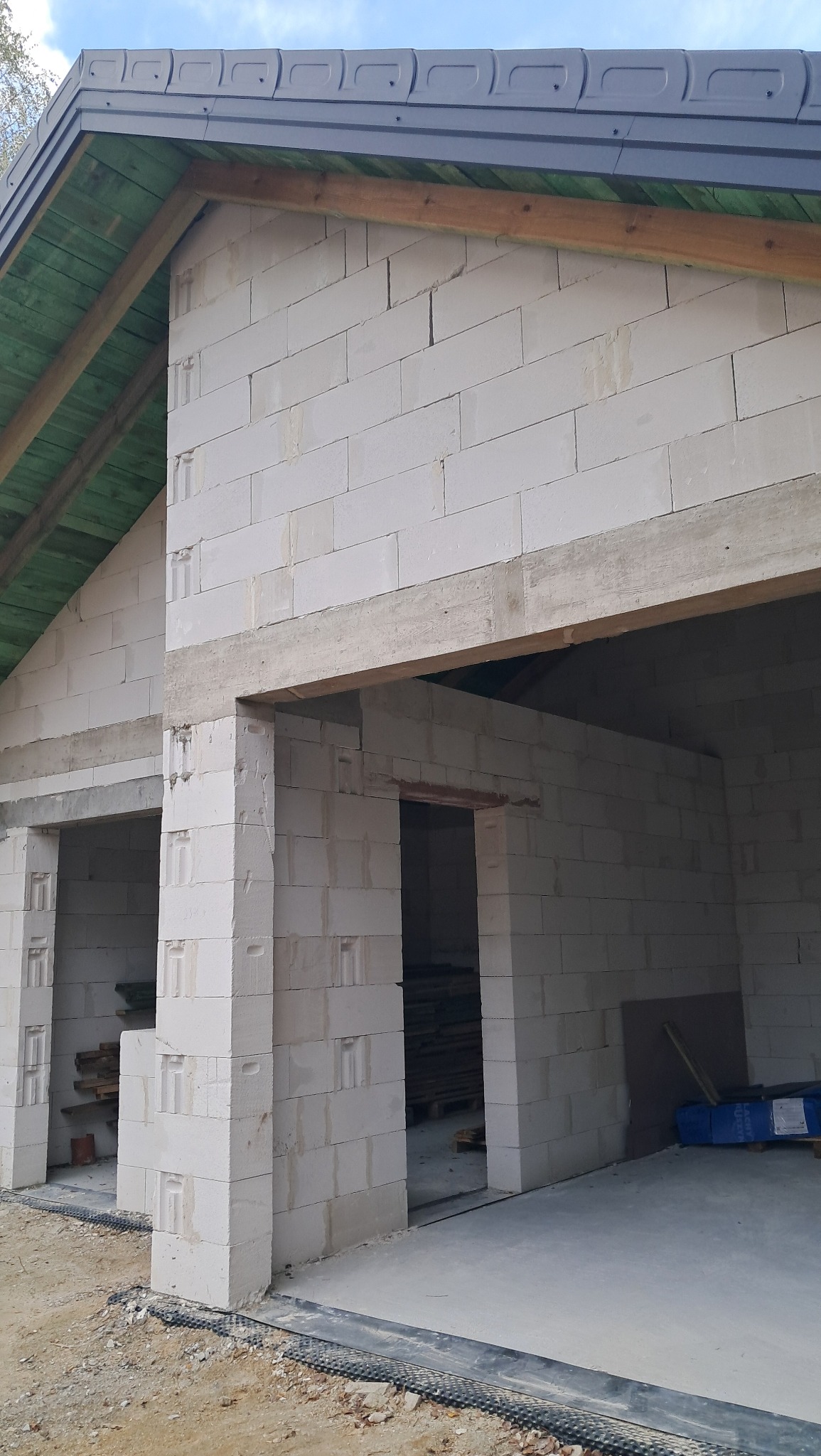 Surowy stan domu z białych bloczków, widoczne otwory okienne i drzwiowe, drewniana konstrukcja dachu pod szarą blachodachówką, widoczne materiały budowlane wewnątrz.