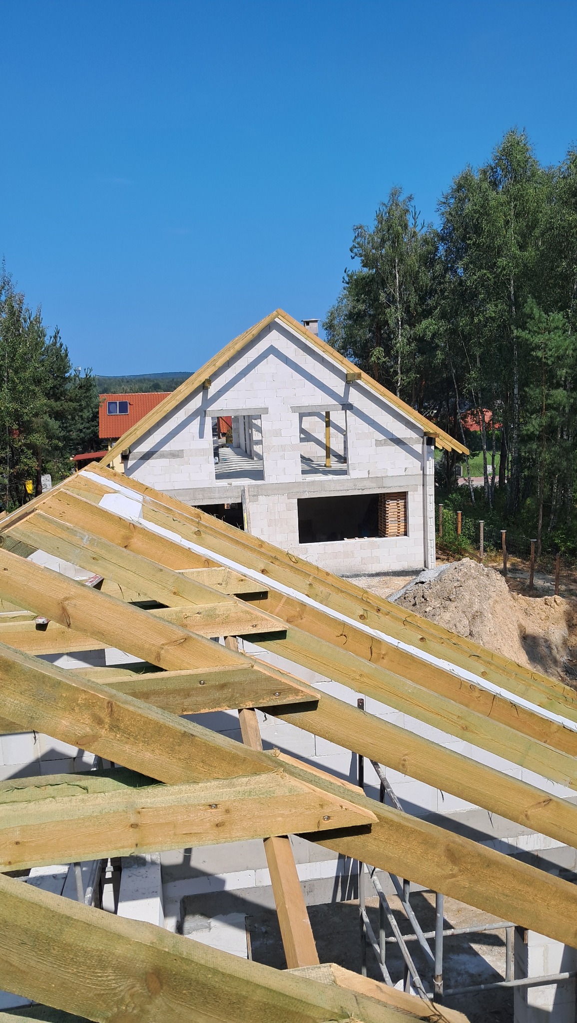 Widok na budowę domu murowanego z perspektywy drewnianej konstrukcji dachu, w tle widać las i błękitne niebo.