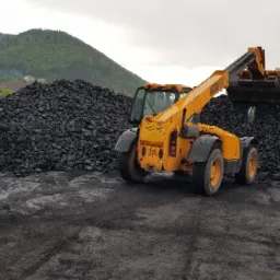 Żółta ładowarka teleskopowa JCB nabiera węgiel z pryzmy na placu składowym, w tle zalesione wzgórza.