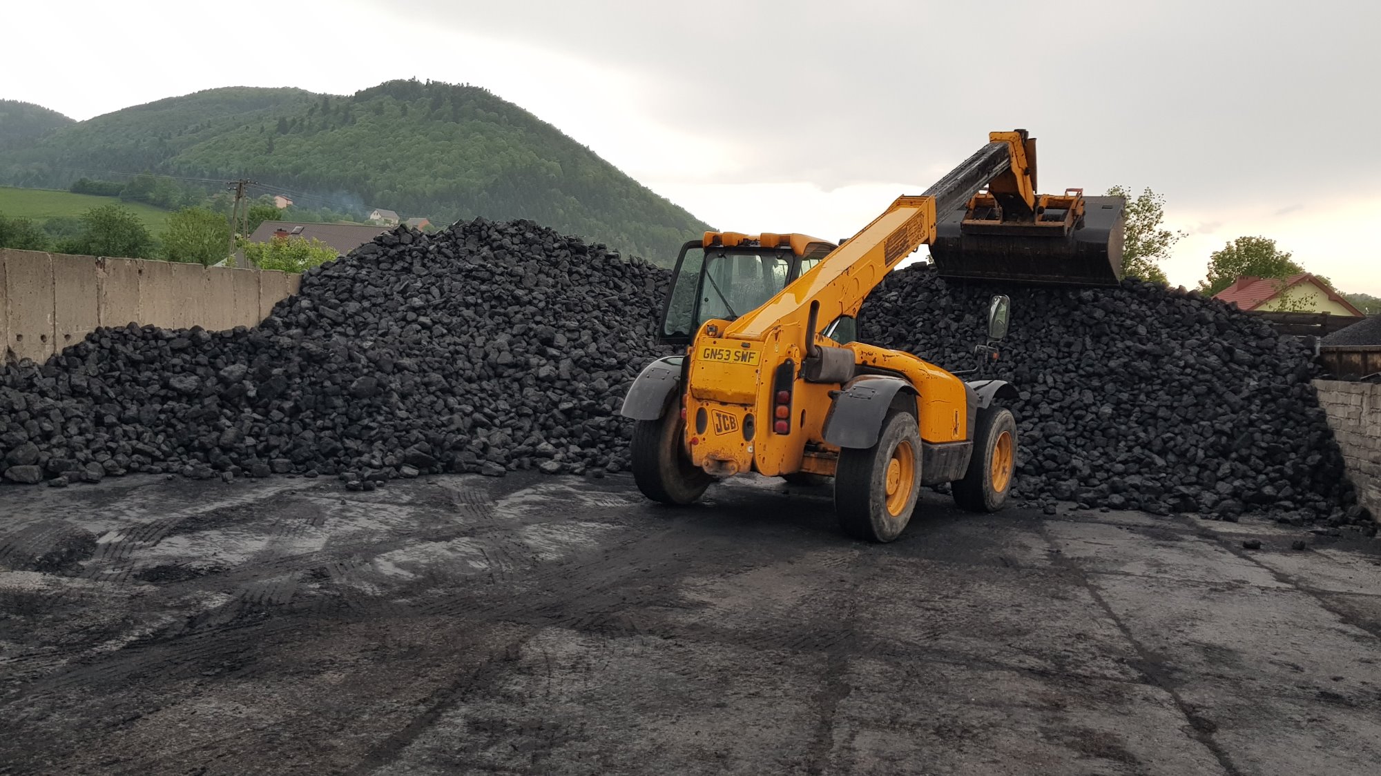 Żółta ładowarka teleskopowa JCB nabiera węgiel z pryzmy na placu składowym, w tle zalesione wzgórza.
