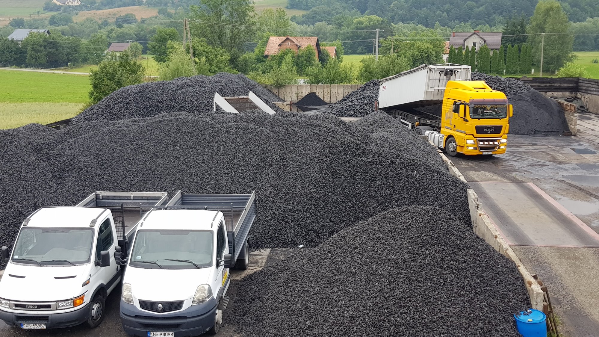 Góra ekogroszku na składzie opału, żółta ciężarówka MAN w tle, dwa białe busy z otwartymi skrzyniami ładunkowymi na pierwszym planie, zieleń drzew i budynki w oddali.