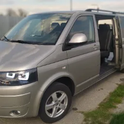 Szary Volkswagen Transporter T5 z otwartymi drzwiami bocznymi, widoczne czarne fotele z jasnymi przeszyciami, na zewnątrz asfaltowa droga i metalowe ogrodzenie.