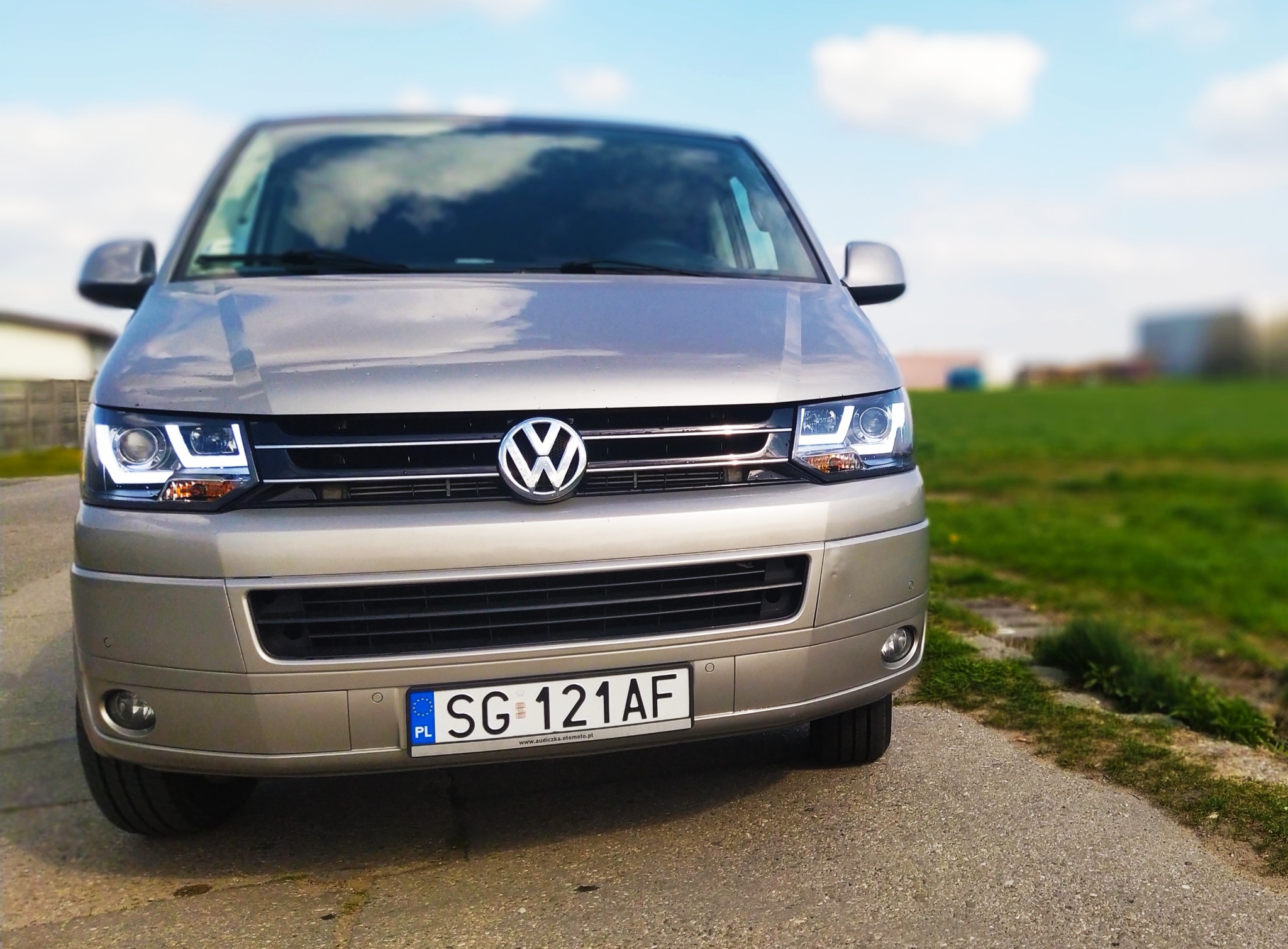 Przód srebrnego busa Volkswagen Transporter z włączonymi światłami LED, zaparkowanego na betonowej nawierzchni, z zielonym polem i budynkiem w tle.