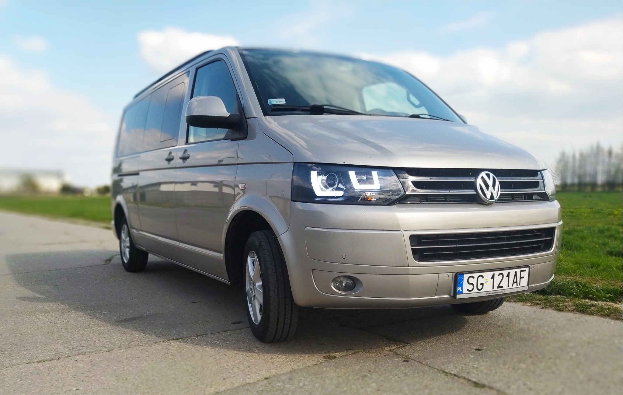 Szary Volkswagen Transporter T6 z włączonymi światłami LED do jazdy dziennej, zaparkowany na asfalcie z widoczną tablicą rejestracyjną SG 121AF.