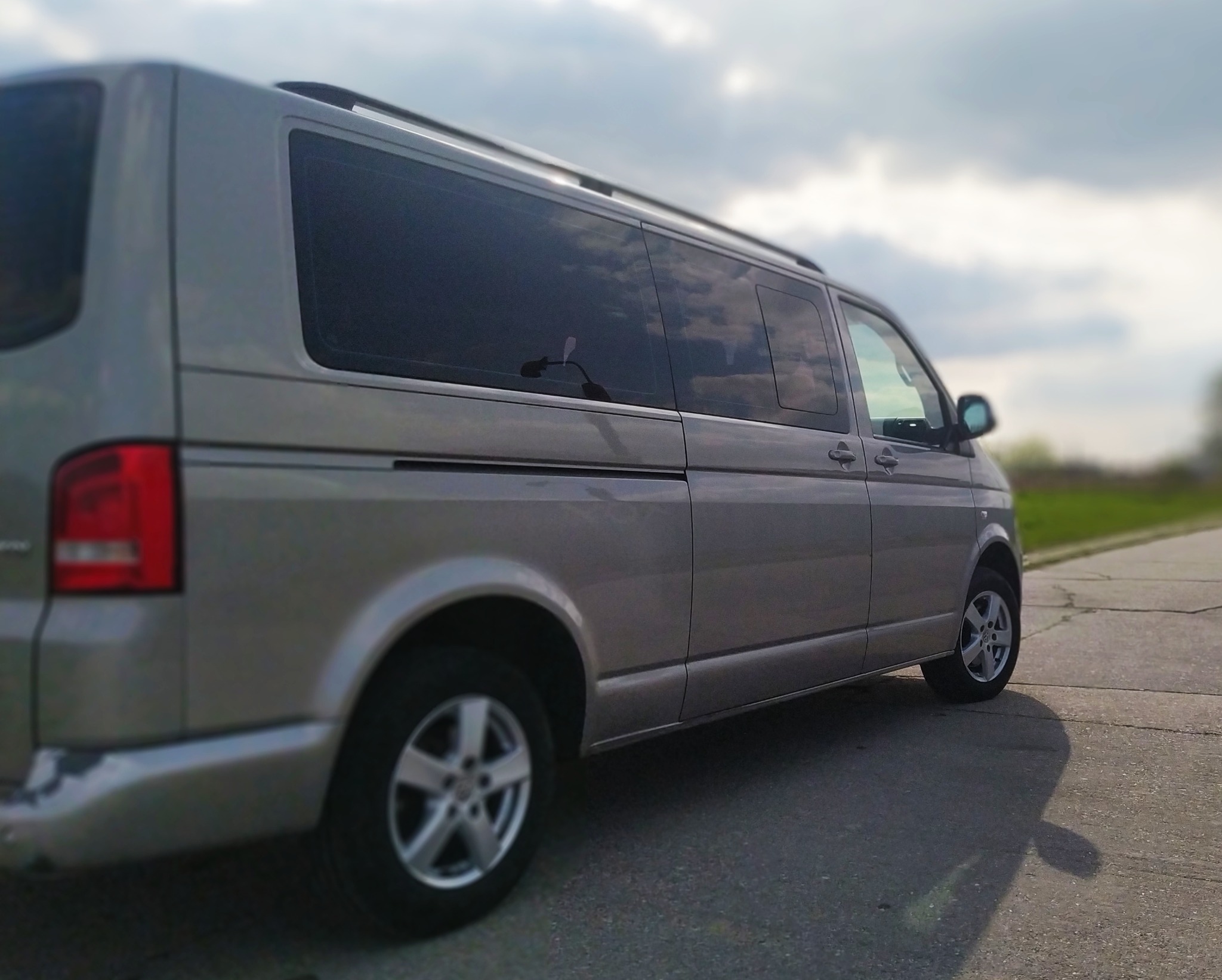 Szary van z relingami dachowymi na betonowej nawierzchni, odbicie chmur w przyciemnionych szybach.