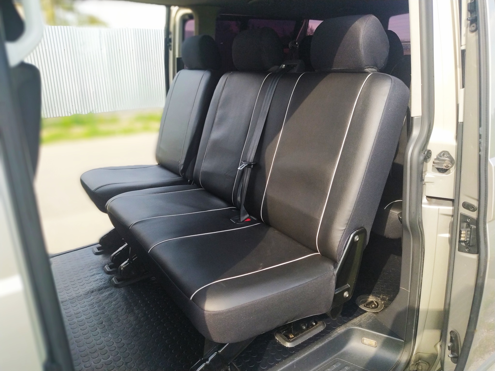 Trzy czarne fotele samochodowe z białymi lamówkami w jasnym vanie, widoczna gumowa podłoga z okrągłym wzorem i szara ściana z blachy falistej za oknem.