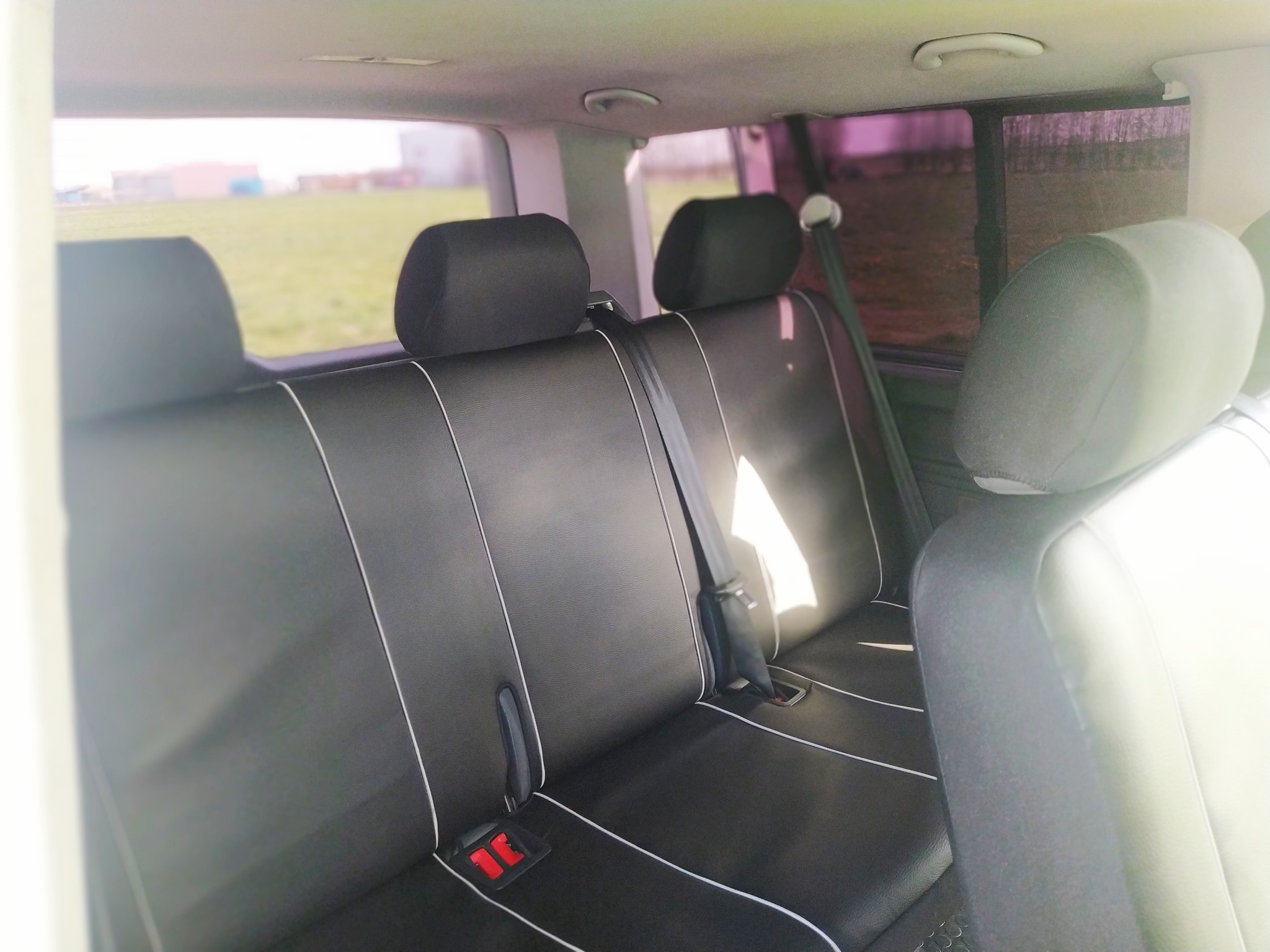 Wnętrze busa z czarną tapicerką foteli z białymi przeszyciami, widok na tylne siedzenia i pasy bezpieczeństwa, za oknem zielona przestrzeń.