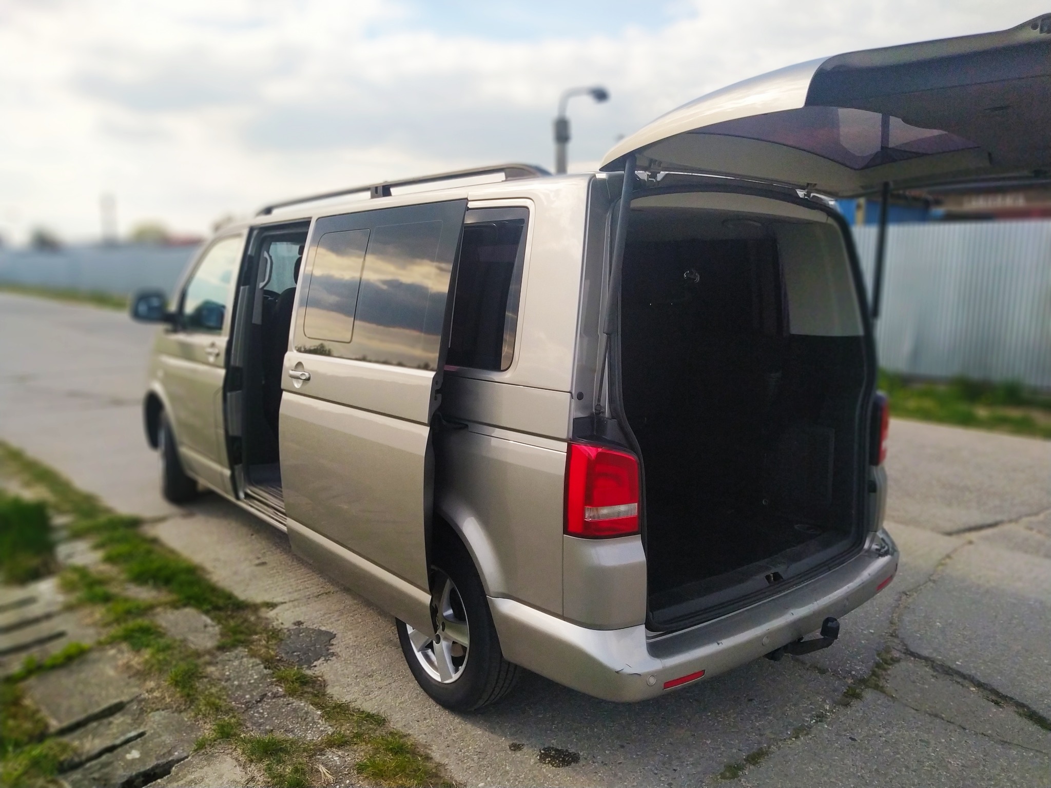Srebrny van z otwartymi drzwiami bocznymi i podniesioną klapą bagażnika, widok z tyłu na ulicy.