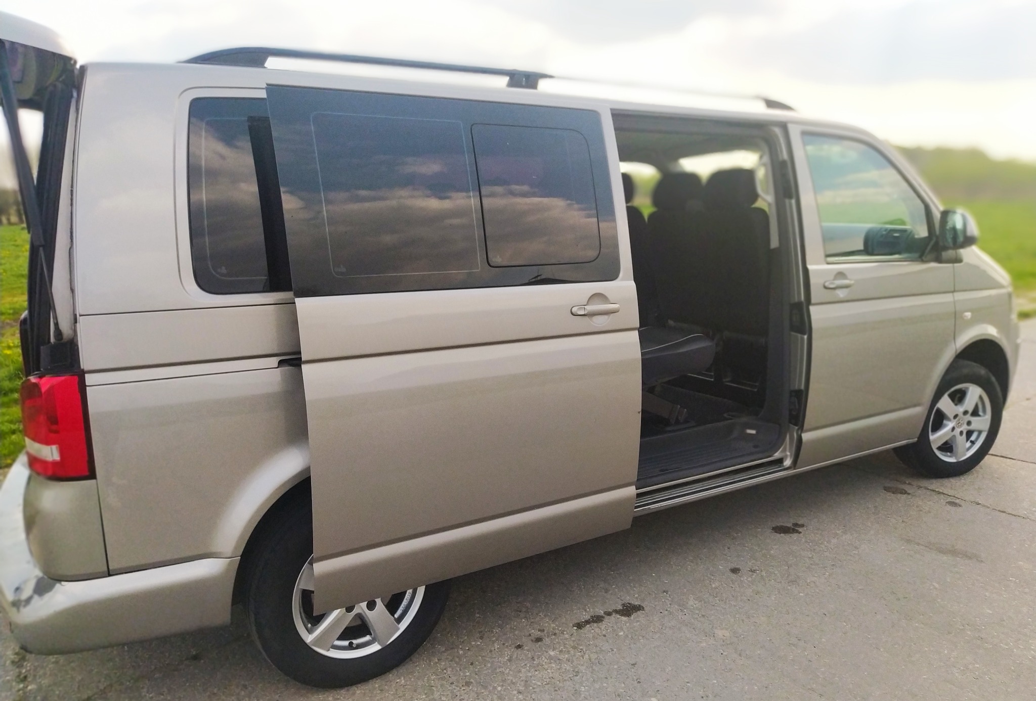 Beżowy van z otwartymi drzwiami bocznymi i tylnymi, widoczne siedzenia wewnątrz, relingi dachowe.