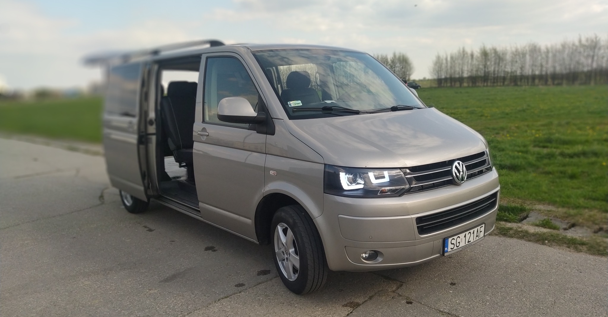 Jasnoszary Volkswagen Transporter z otwartymi drzwiami przesuwnymi, zaparkowany na asfaltowej drodze z polnym tłem.