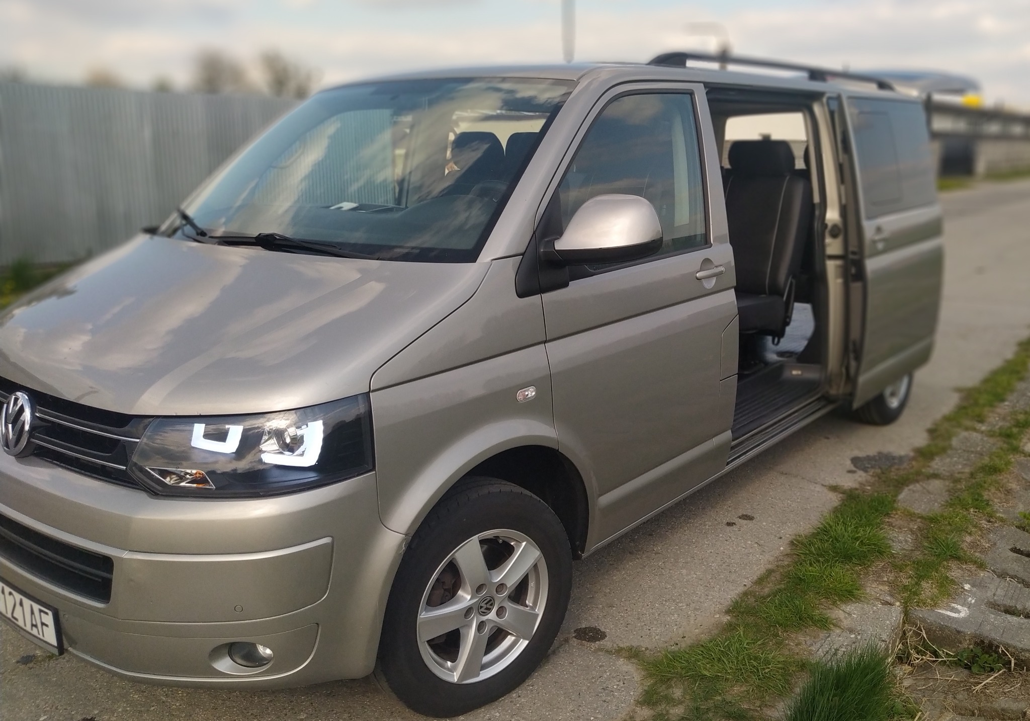 Szary Volkswagen Transporter T5 z otwartymi drzwiami bocznymi, widoczne czarne fotele z jasnymi przeszyciami, na zewnątrz asfaltowa droga i metalowe ogrodzenie.