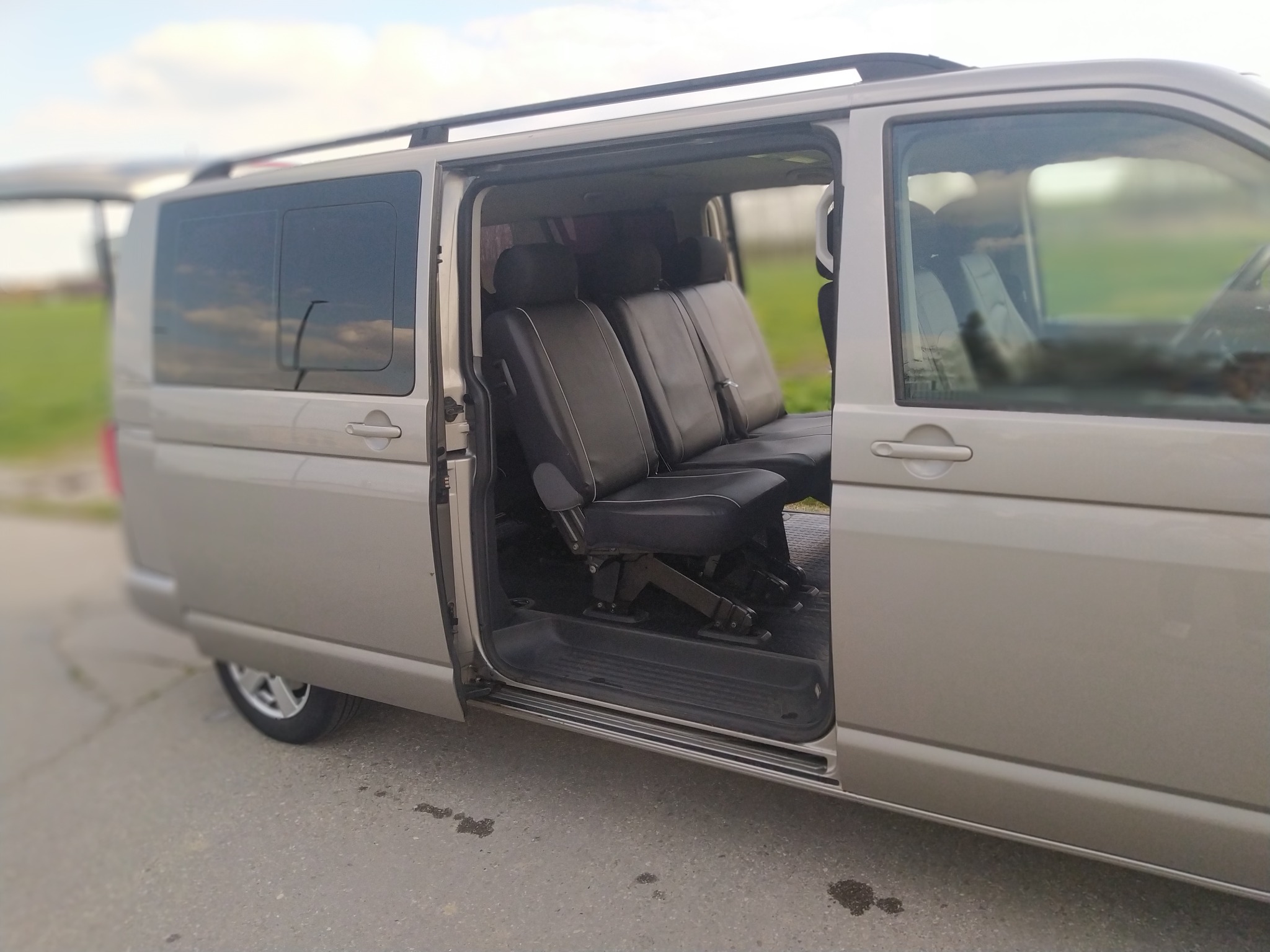 Jasnoszary van z otwartymi drzwiami bocznymi, ukazujący czarne skórzane siedzenia pasażerskie z regulowanymi zagłówkami i mechanizmem składania, na zewnątrz widoczne zielone pole i pochmurne niebo.