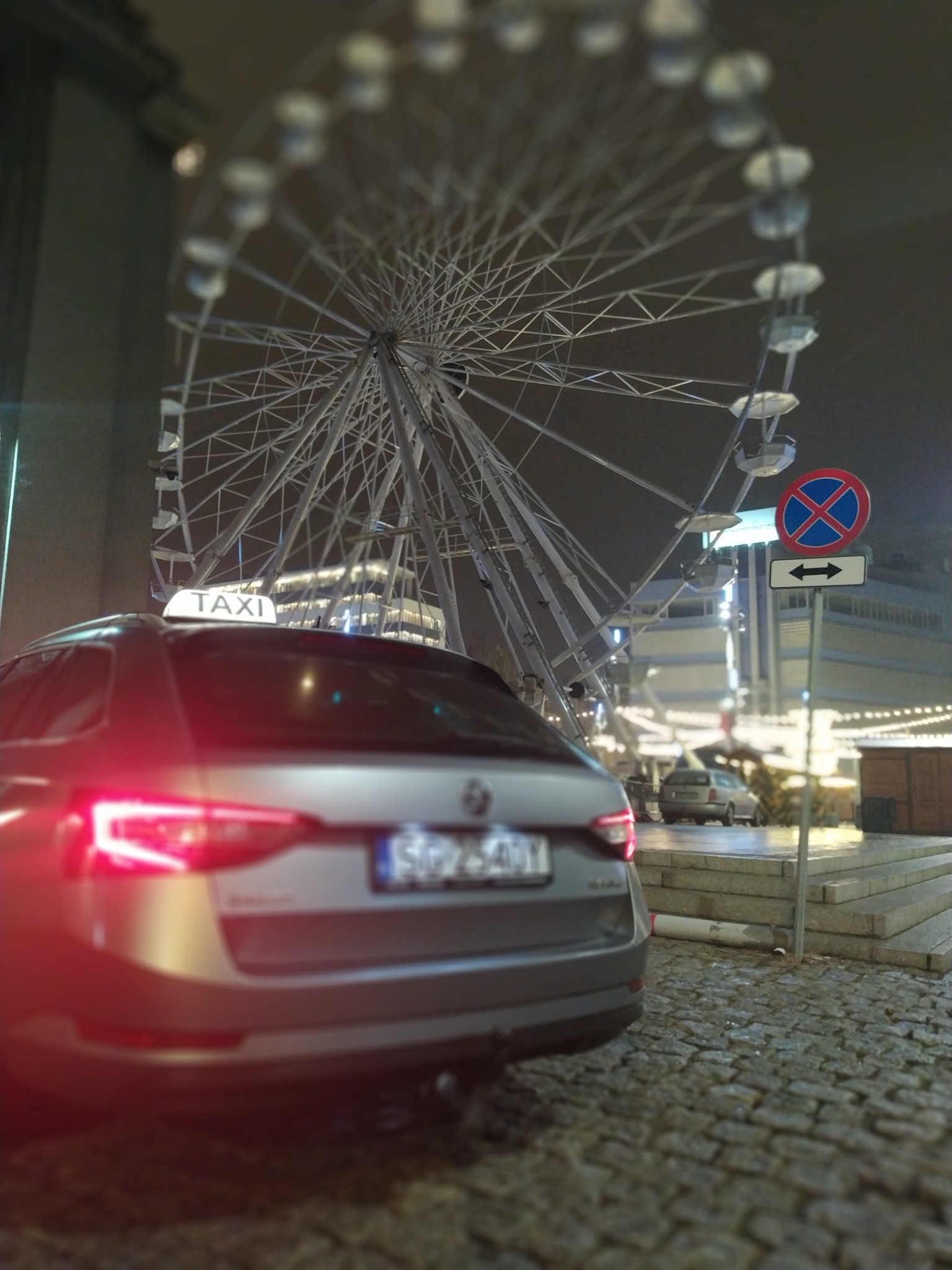 Szary samochód typu kombi z napisem TAXI na dachu, stojący na brukowanej ulicy nocą, z rozmytym diabelskim młynem w tle.