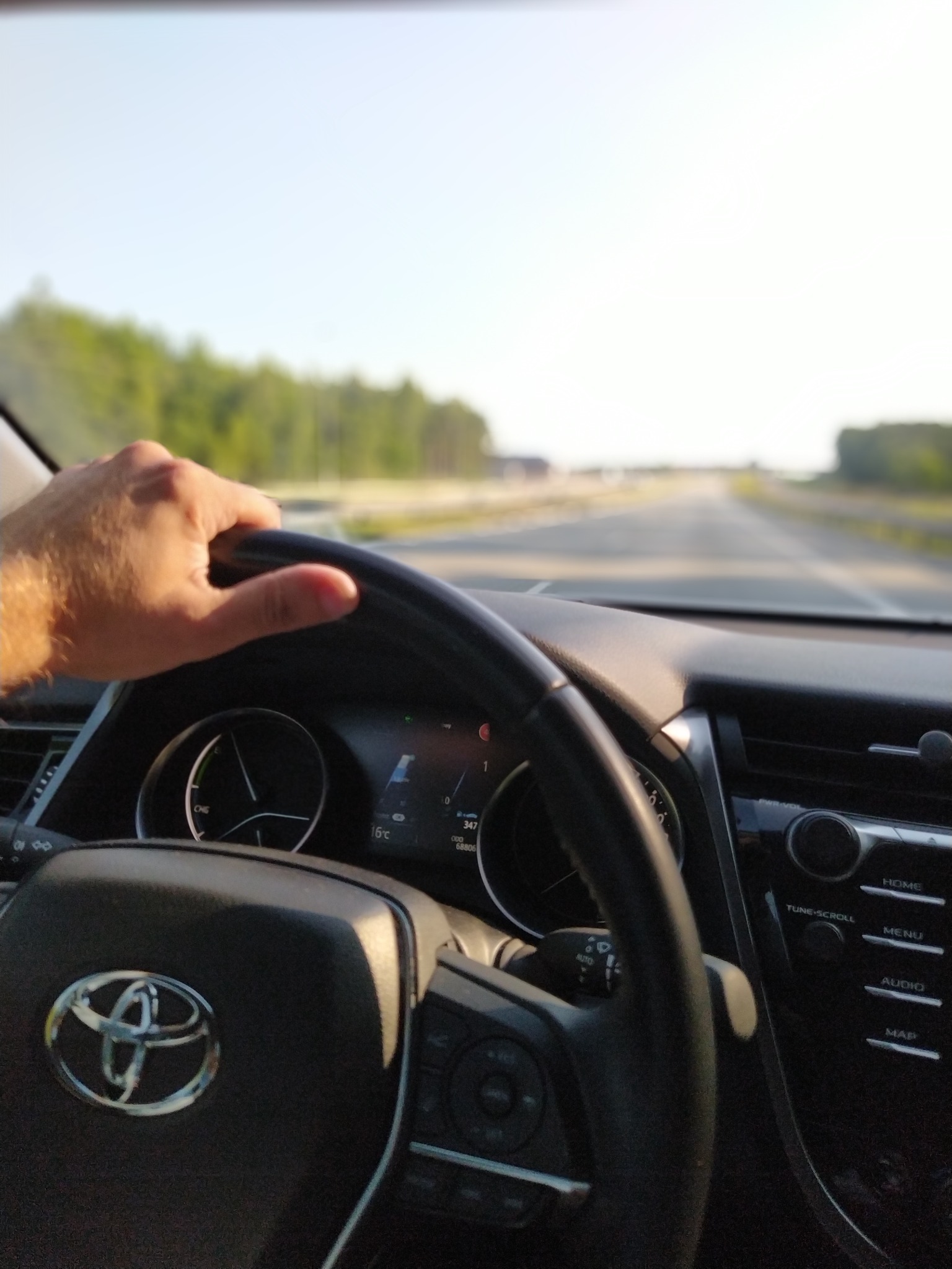 Wnętrze samochodu Toyota z widokiem na drogę z perspektywy kierowcy, ręka na kierownicy, deska rozdzielcza z wyświetlaczem i logo marki.