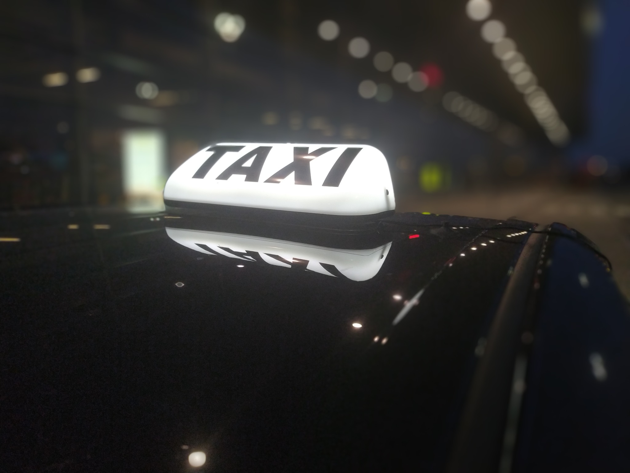Świecący szyld TAXI na dachu ciemnego pojazdu, nocne oświetlenie w tle.