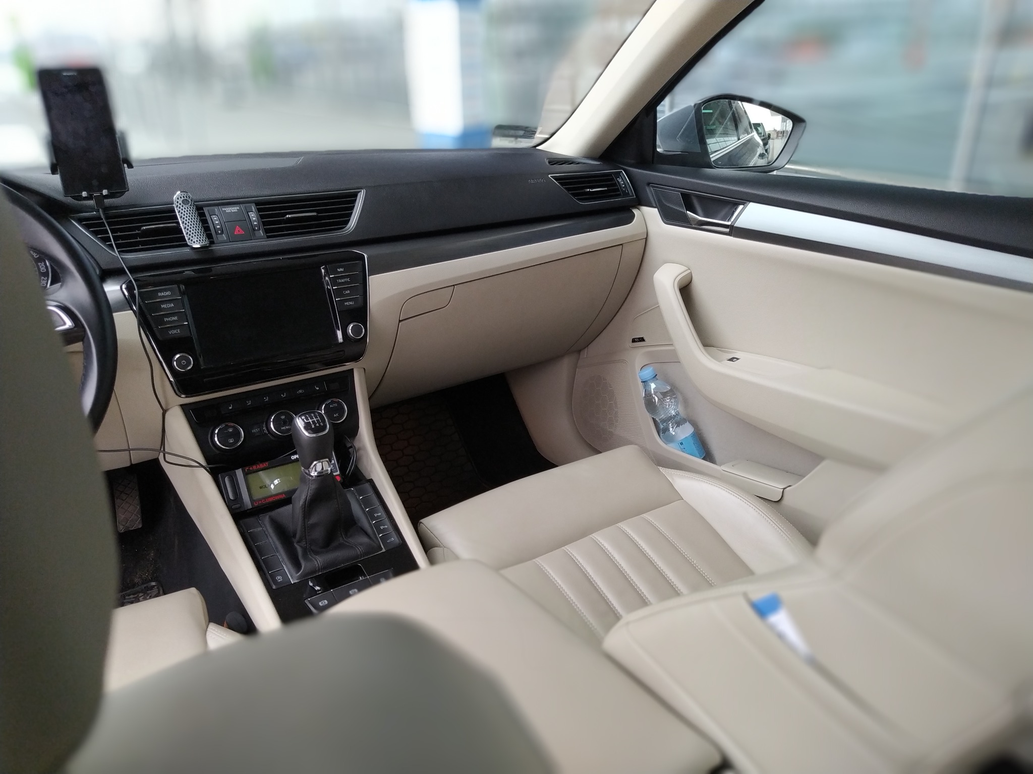 Wnętrze samochodu osobowego z jasną tapicerką skórzaną, widok z tylnego siedzenia na deskę rozdzielczą, uchwyt na telefon i butelkę wody w drzwiach.