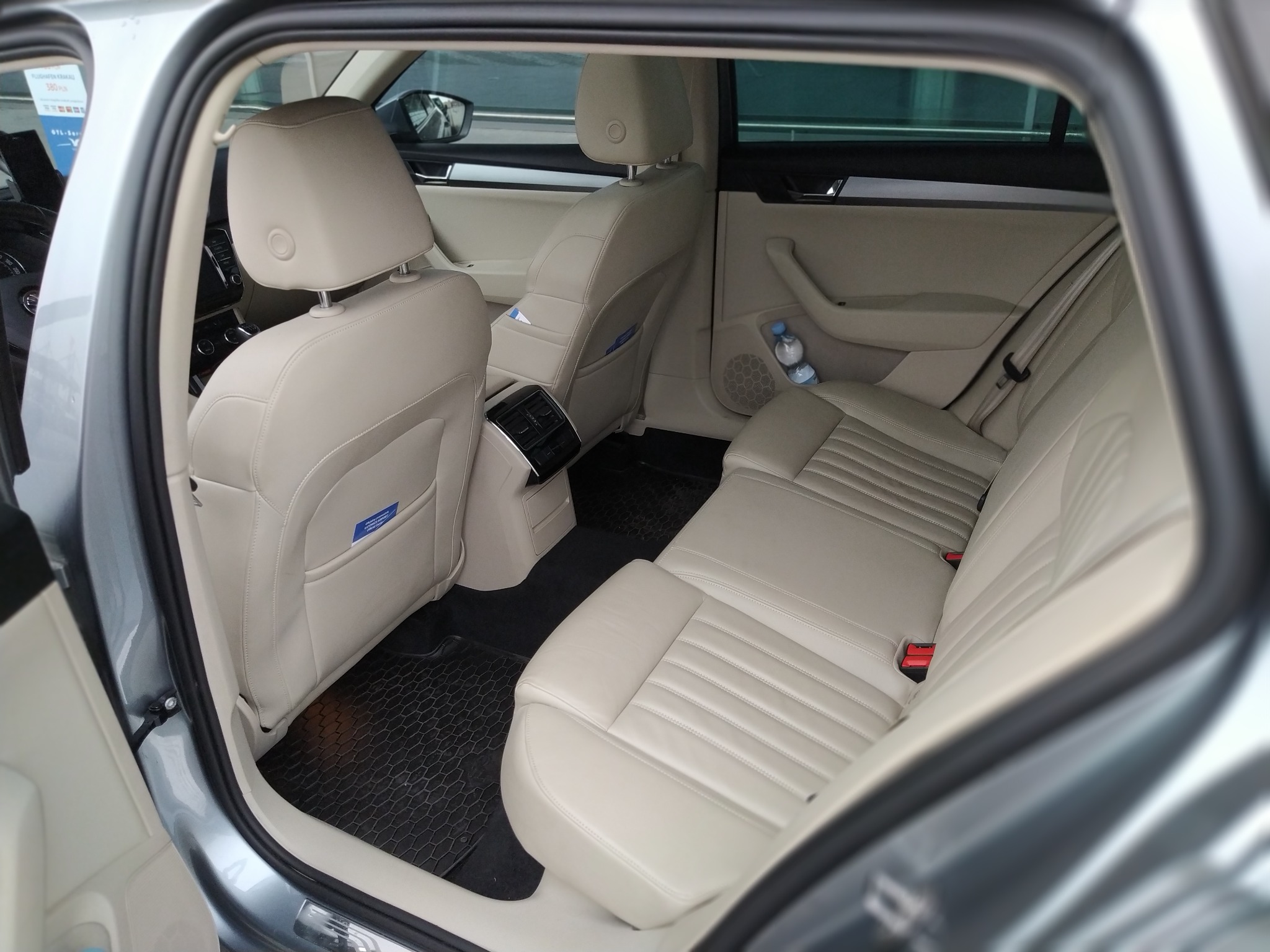 Wnętrze samochodu z jasną skórzaną tapicerką, widok na tylne siedzenia i czarne dywaniki, butelka wody w kieszeni fotela.