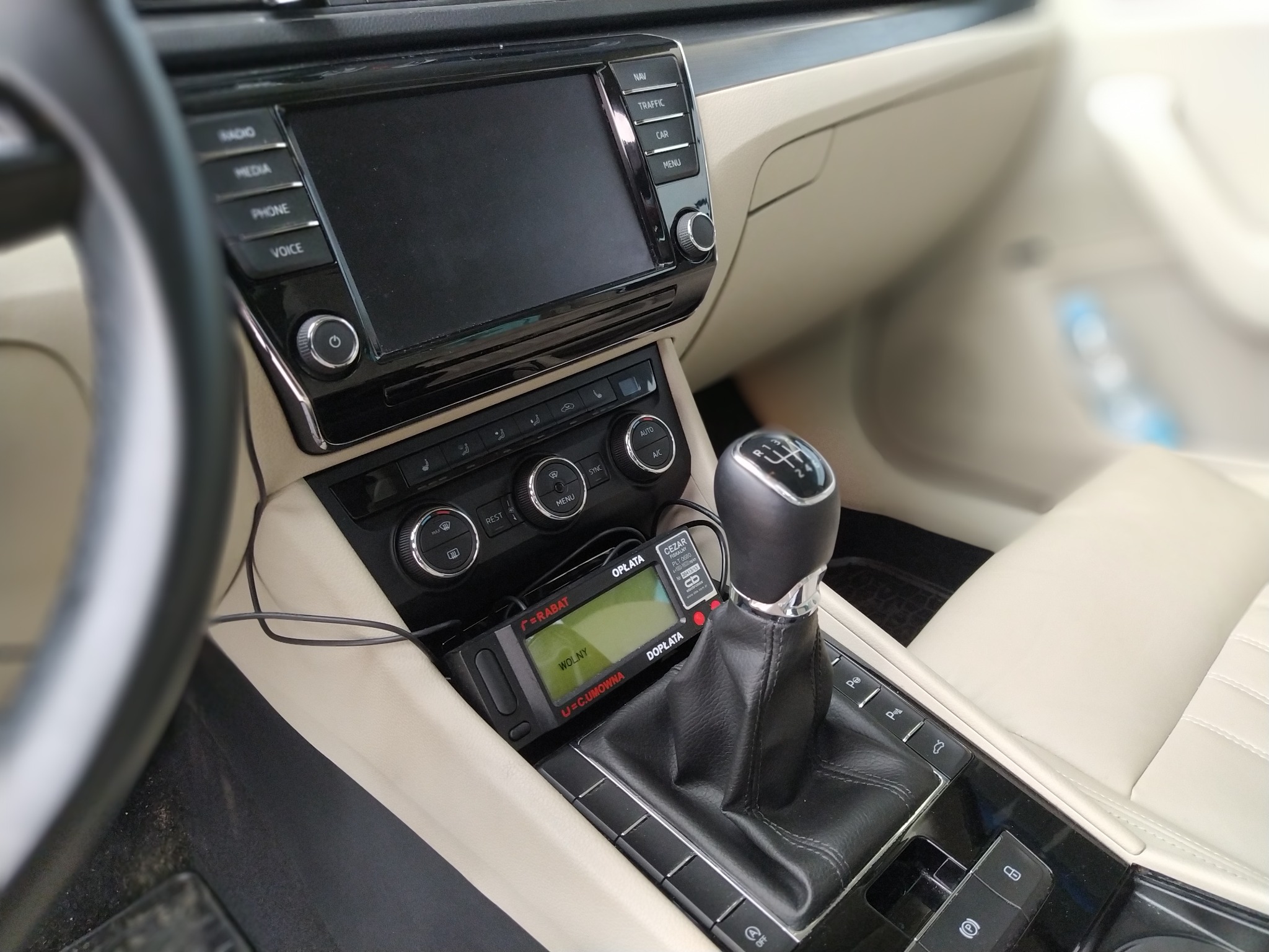 Wnętrze samochodu z zamontowanym taksometrem 'OPŁATA' obok dźwigni zmiany biegów, widoczny panel sterowania klimatyzacją i systemem multimedialnym.
