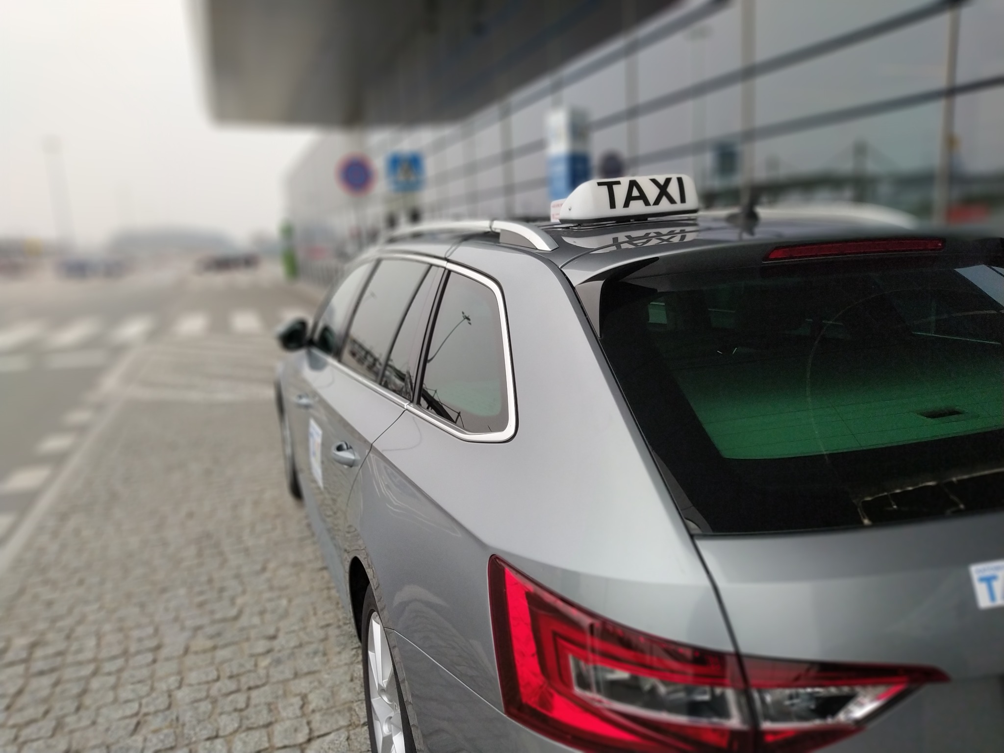 Szary samochód typu kombi z napisem TAXI na dachu, zaparkowany na zewnątrz budynku z przeszkloną fasadą.