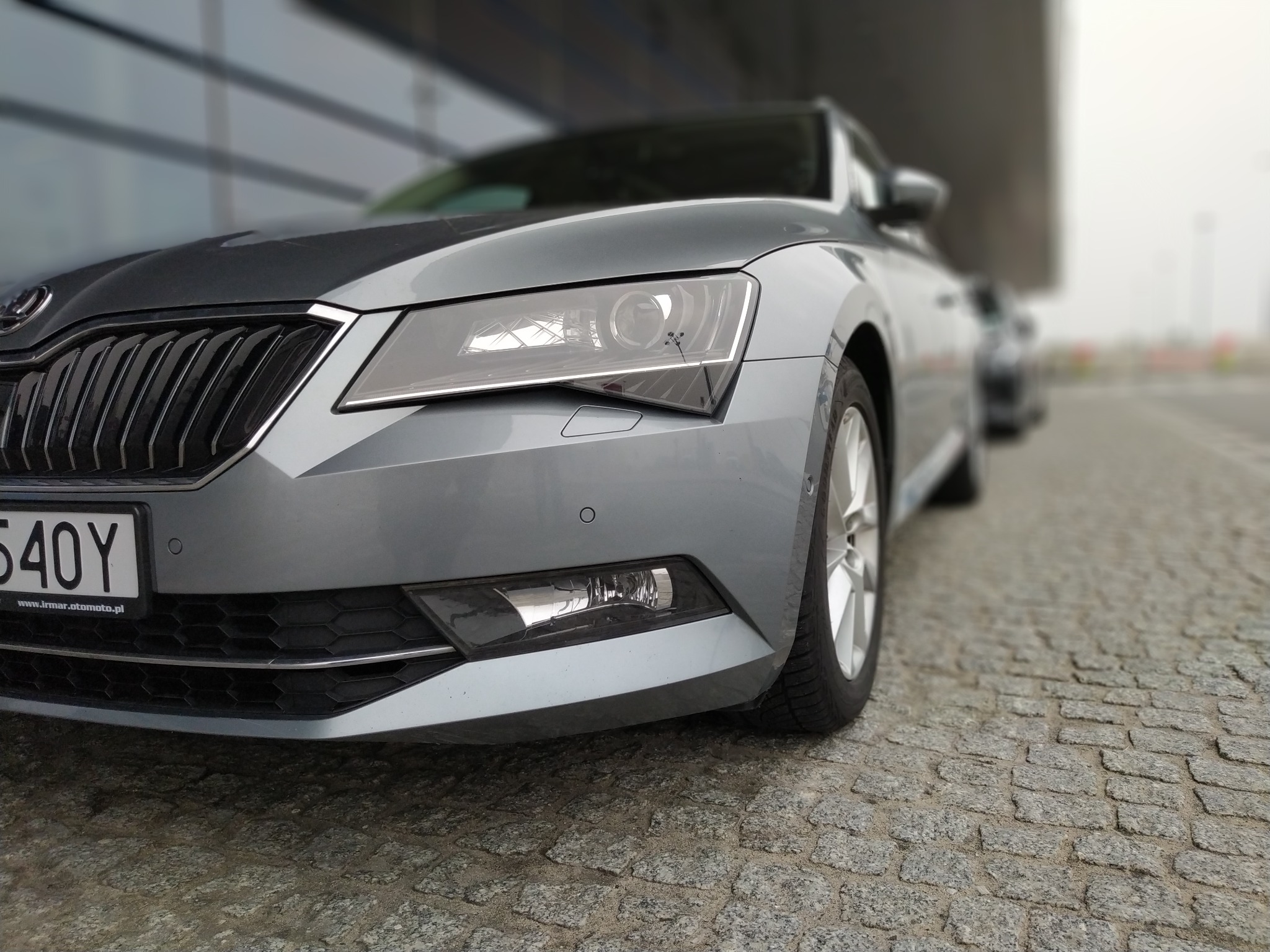 Przód szarej Skody Superb z widoczną tablicą rejestracyjną na brukowanej nawierzchni, nowoczesny budynek w tle.