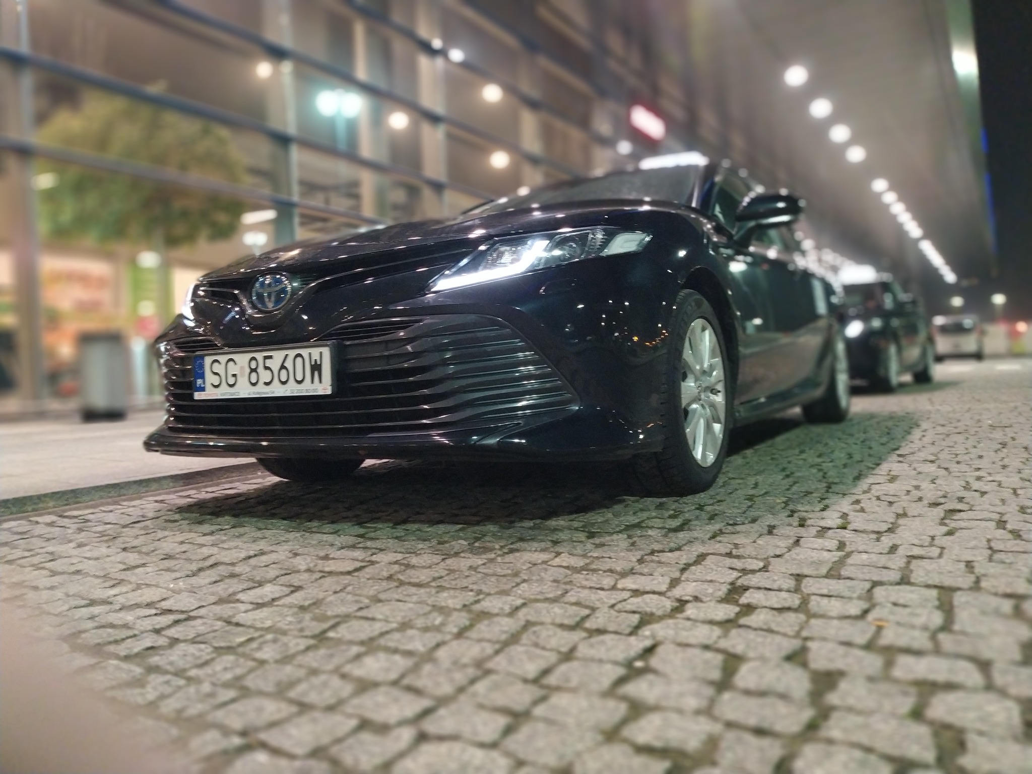 Czarny samochód osobowy z włączonymi światłami, zaparkowany na brukowanej ulicy, z widoczną tablicą rejestracyjną SG 8560W i innymi autami w tle.