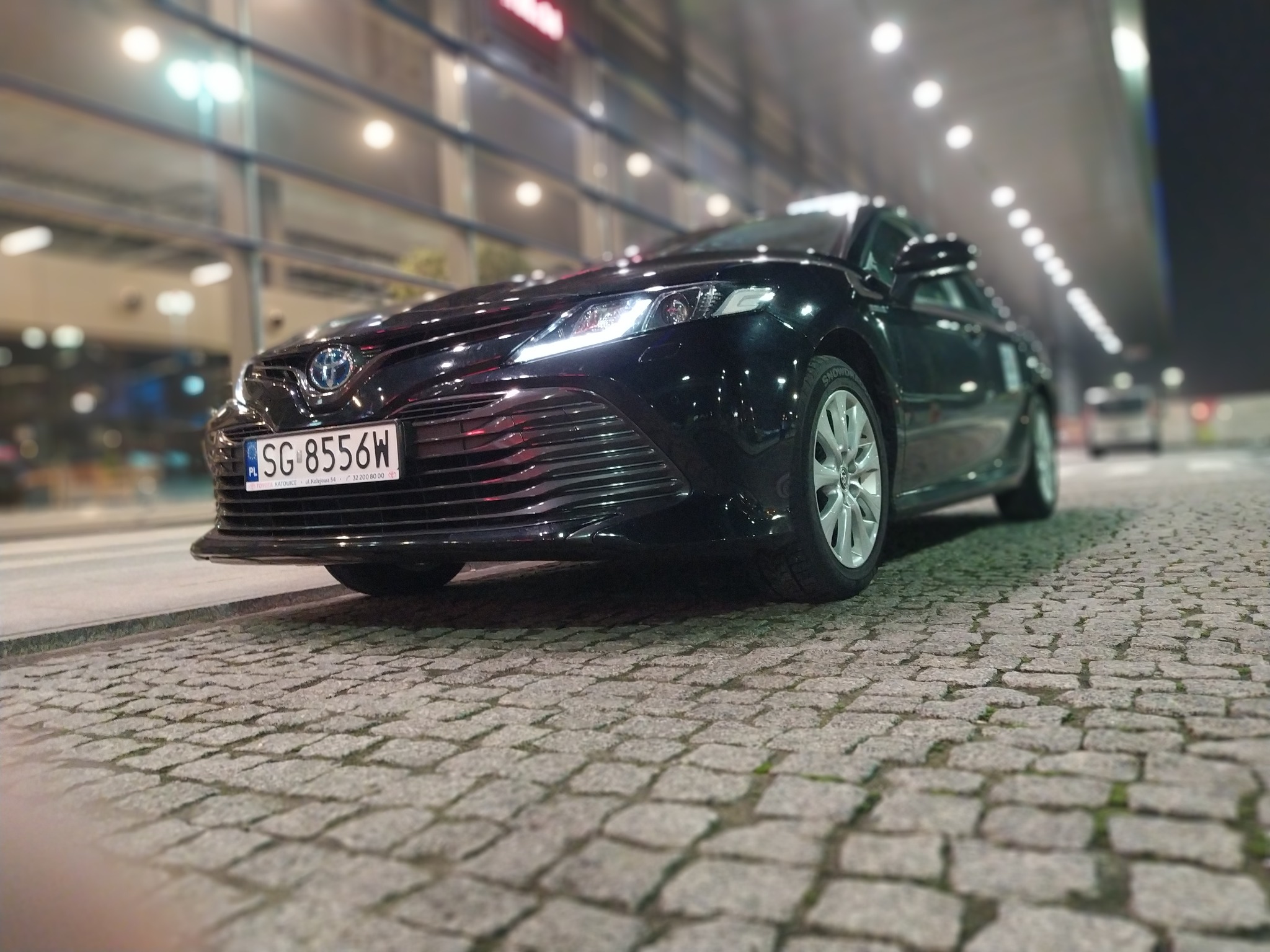 Czarny samochód osobowy Toyota Camry zaparkowany na brukowanej ulicy, w tle budynek z przeszkloną fasadą i oświetleniem.