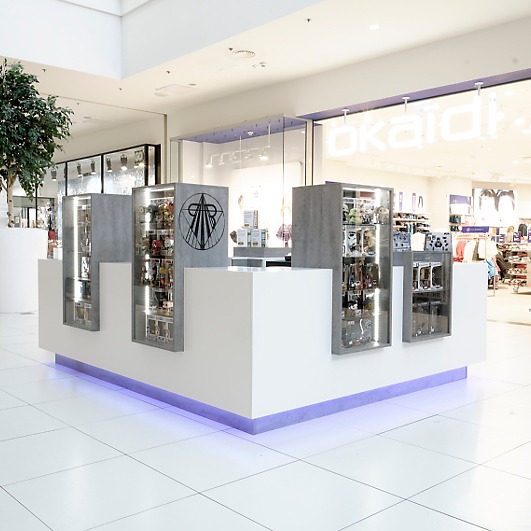 Biały kiosk handlowy z szarymi elementami i wbudowanymi witrynami w centrum handlowym, podświetlony fioletowym światłem od dołu.