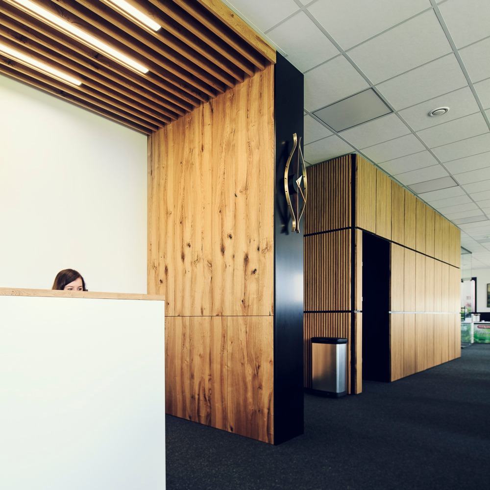 Recepcja biurowa z drewnianymi panelami ściennymi i sufitem z listew, widoczna osoba za ladą, minimalistyczny design z oświetleniem LED.