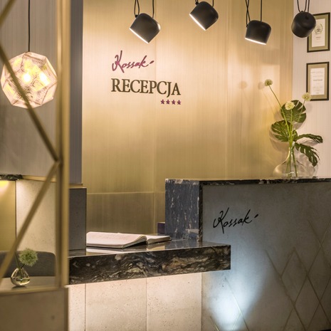 Recepcja hotelu Kossak z marmurowym blatem, oświetleniem punktowym i dekoracyjną lampą geometryczną, widok zza metalowej konstrukcji.