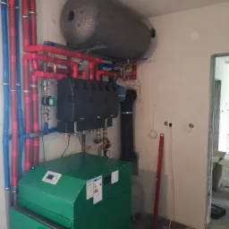 Kotłownia w trakcie montażu: zielony piec, czarny podajnik, szary zbiornik akumulacyjny. Widoczne rury w izolacji czerwonej i niebieskiej oraz poziomica oparta o ścianę.