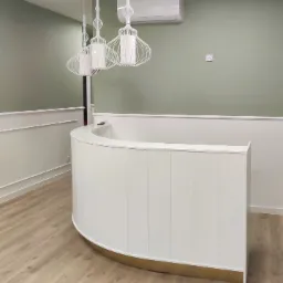 Nowoczesne wnętrze biura z zaokrągloną białą recepcją z złotą podstawą, trzema wiszącymi lampami z białymi abażurami i klimatyzacją na pistacjowej ścianie.