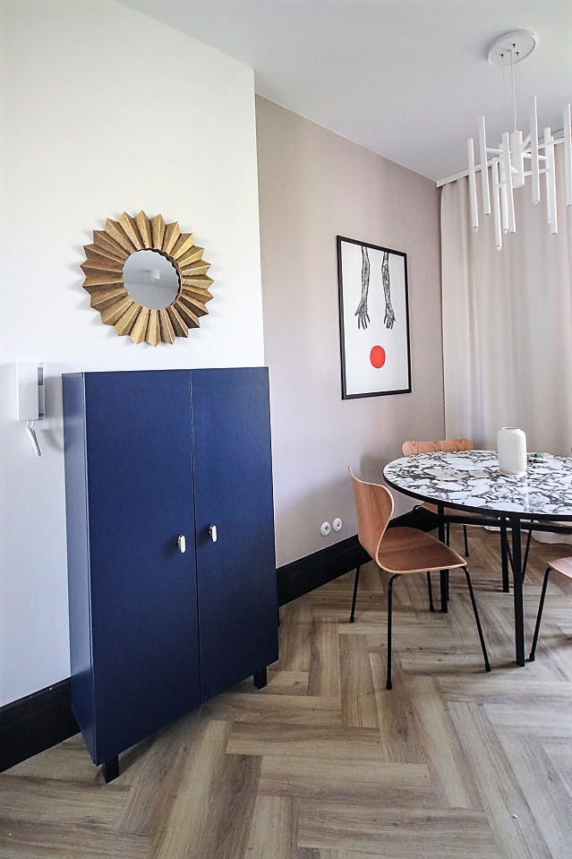 Pokój z granatową szafką, lustrem w złotej ramie, okrągłym stołem z marmurowym blatem i parkietem ułożonym w jodełkę.