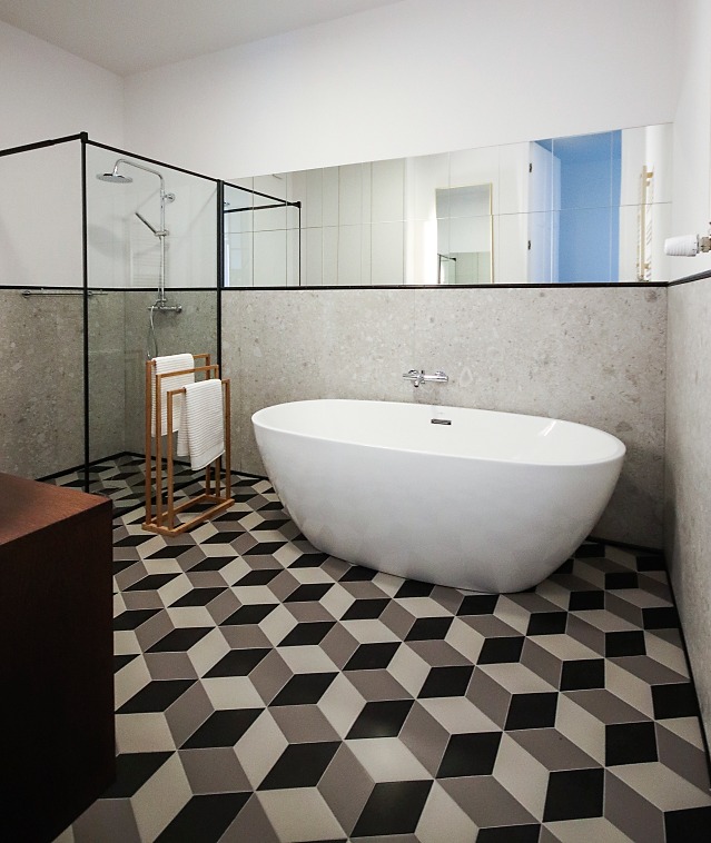 Nowoczesna łazienka z wolnostojącą wanną, kabiną prysznicową z czarnymi ramami i podłogą w geometryczne wzory 3D, uzupełniona o drewniany wieszak na ręczniki.