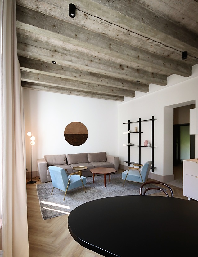 Jasne wnętrze salonu z betonowym stropem belkowym, parkietem w jodełkę, beżową sofą i błękitnymi fotelami.