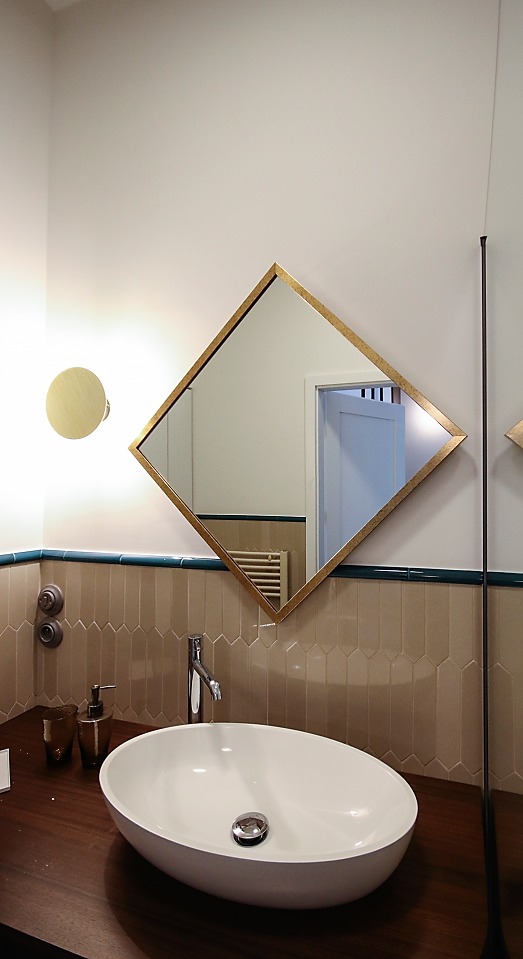 Owalna umywalka nablatowa z chromowaną baterią i dozownikiem mydła, na ciemnym blacie w łazience z beżowymi płytkami i lustrem w złotej ramie w kształcie rombu.