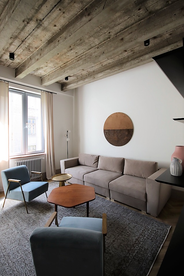Jasne, przestronne wnętrze mieszkania z betonowym sufitem, szarą sofą modułową, błękitnymi fotelami i dywanem w stylu vintage.