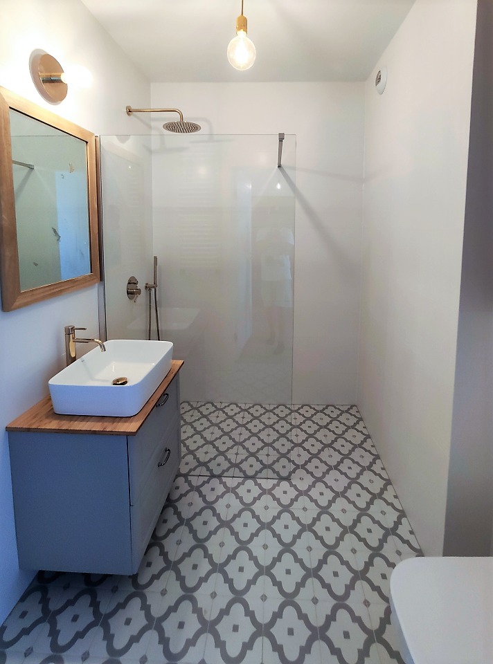 Nowoczesna łazienka z kabiną prysznicową typu walk-in, umywalką nablatową na szafce w kolorze gołębim i wzorzystą szaro-białą podłogą. Widoczna złota armatura i oświetlenie punktowe.