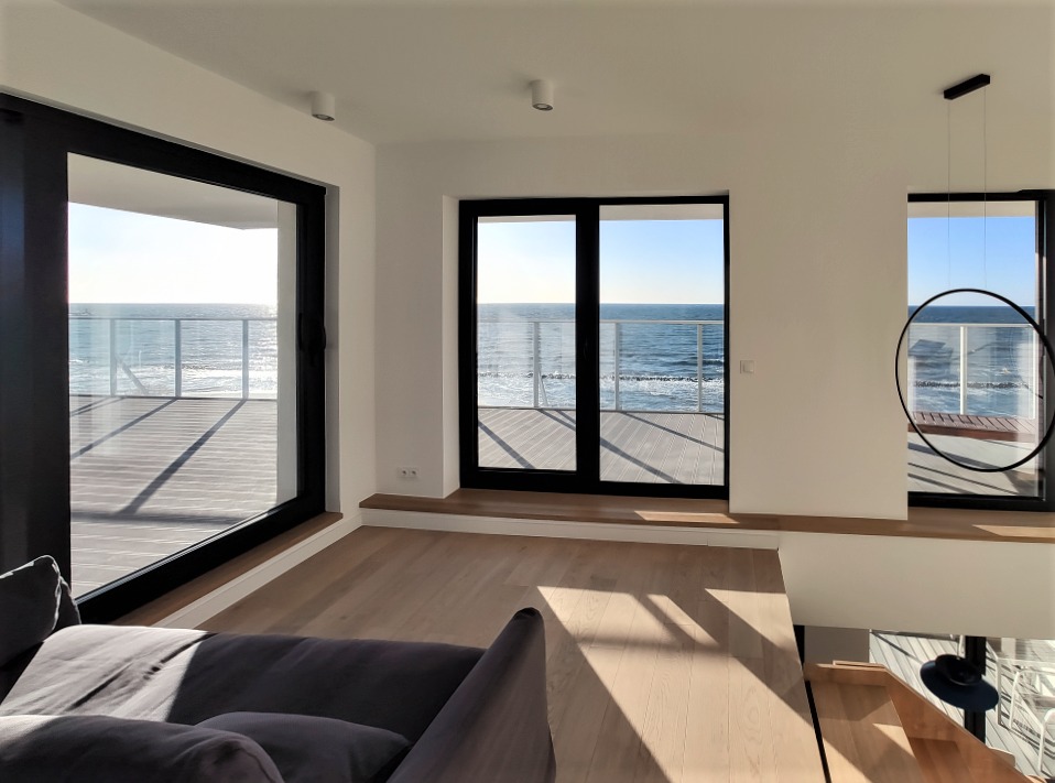 Jasne, przestronne wnętrze z podłogą z jasnego drewna i widokiem na morze przez duże okna balkonowe z czarnymi ramami, częściowo widoczna szara sofa w lewym dolnym rogu.