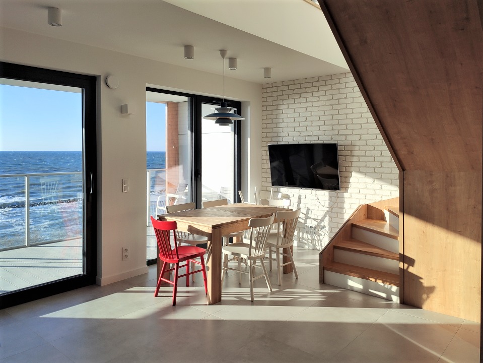 Jasne wnętrze jadalni z widokiem na morze, drewniany stół z krzesłami, czerwone krzesło wyróżniające się na tle białych, ściana z białej cegły z telewizorem, drewniane schody, duże okna przesuwne.