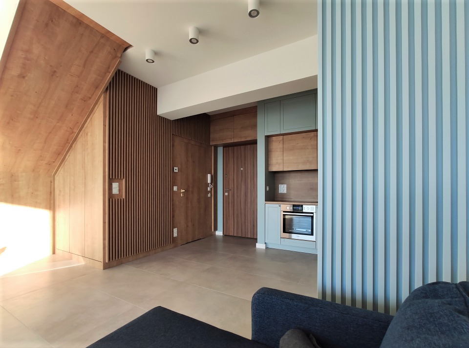 Nowoczesne wnętrze mieszkania z drewnianymi panelami ściennymi, zabudową kuchenną w odcieniach szarości i błękitu, oraz podświetlanymi punktowo sufitami.