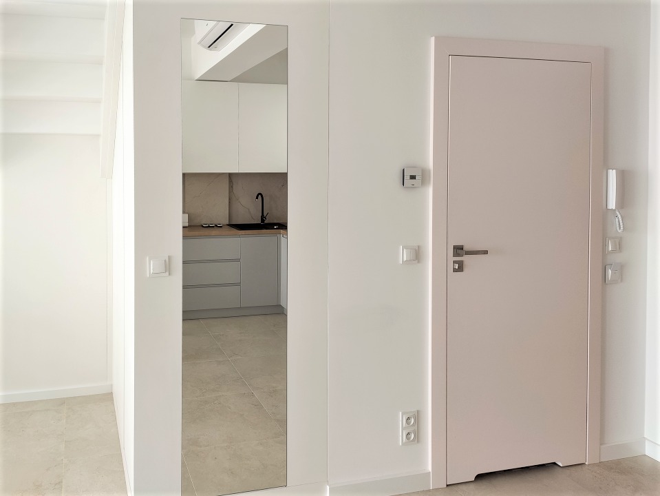 Nowoczesne wnętrze mieszkania: białe ściany, lustro w przedpokoju odbijające widok na minimalistyczną kuchnię z szarymi szafkami i czarną armaturą, beżowa podłoga z płytek, jasnoróżowe drzwi...
