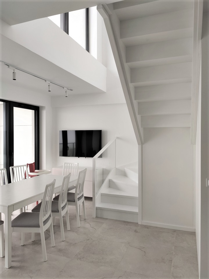 Jasne wnętrze z białymi schodami, stołem i krzesłami w minimalistycznym stylu, oświetlenie szynowe na suficie, telewizor na ścianie, marmurowa podłoga.