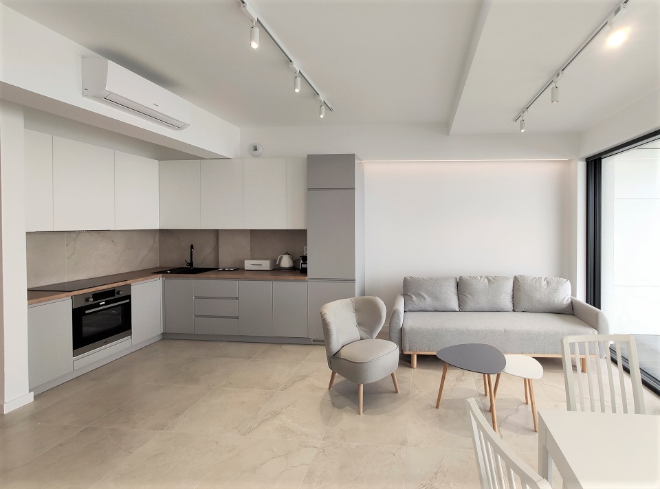 Jasne, nowoczesne wnętrze mieszkania z aneksem kuchennym, szarą zabudową i sprzętami, beżową podłogą z płytek imitujących marmur oraz minimalistycznym salonem z szarą sofą i okrągłymi stolikami.